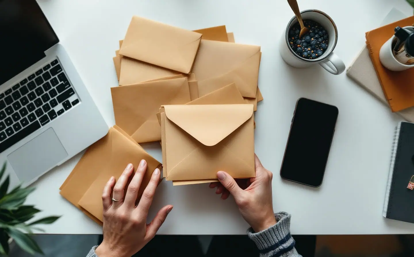 Hands sorting plain envelopes beside a closed laptop and a dark phone on a white desk